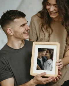 Smiling couple showing a small wooden photo frame with a printed wedding-style photo