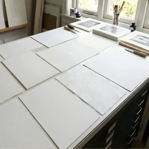 Various textured fine art papers and archival media with different shades of white laid out on a studio table with printmaking tools.