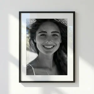 Black framed black and white photos of a smiling woman with freckles wearing a straw hat, hanging on a white wall with natural window light shadows and reflections on the frame's glass.
