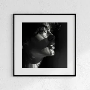 Black framed black and white photograph of a woman's profile looking upward, featuring dramatic striped light and shadow across her face, hanging on a textured white wall.